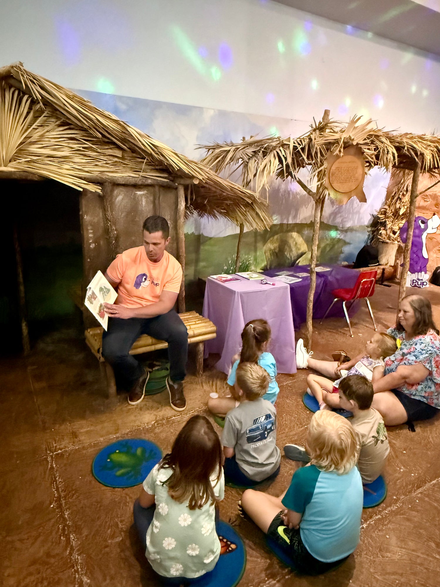 Benjamin A. King reading to children in the Children's Hands-On Museum of Tuscaloosa with straw huts and tables. During storytime Ben was reading his latest picture book Priscilla in the Purple Hood. 