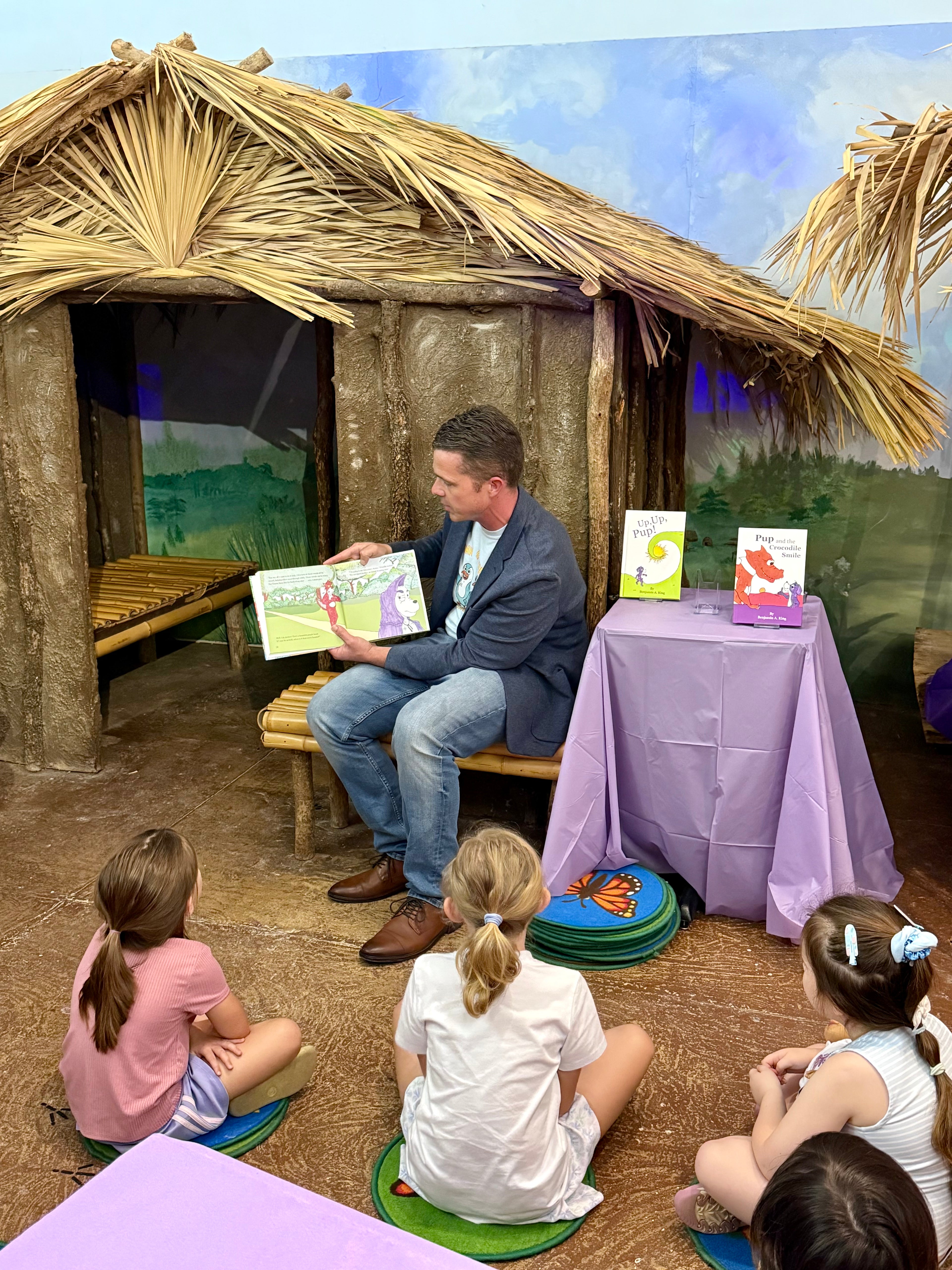 Benjamin A. King reading his Priscilla in the Purple Hood picture book to children in the Children's Hands-On Museum of Tuscaloosa with a rustic setting with straw roof and wooden benches.