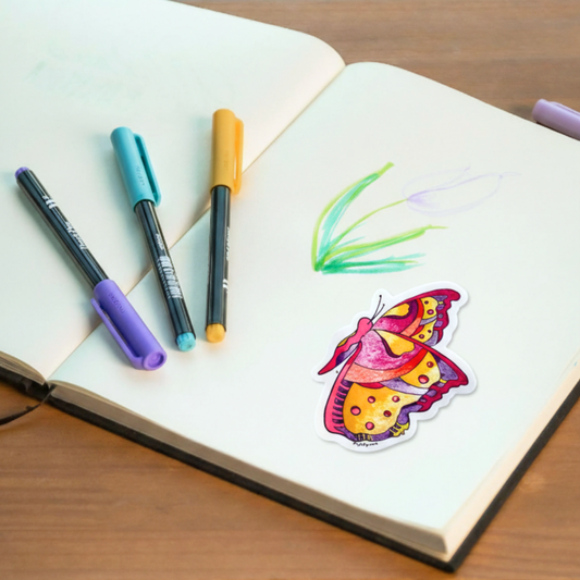 Colorful butterfly sticker placed close to a flower drawing on a notebook with markers on a wooden surface. A butterfly inspired by the world of Prpl Up Pup.