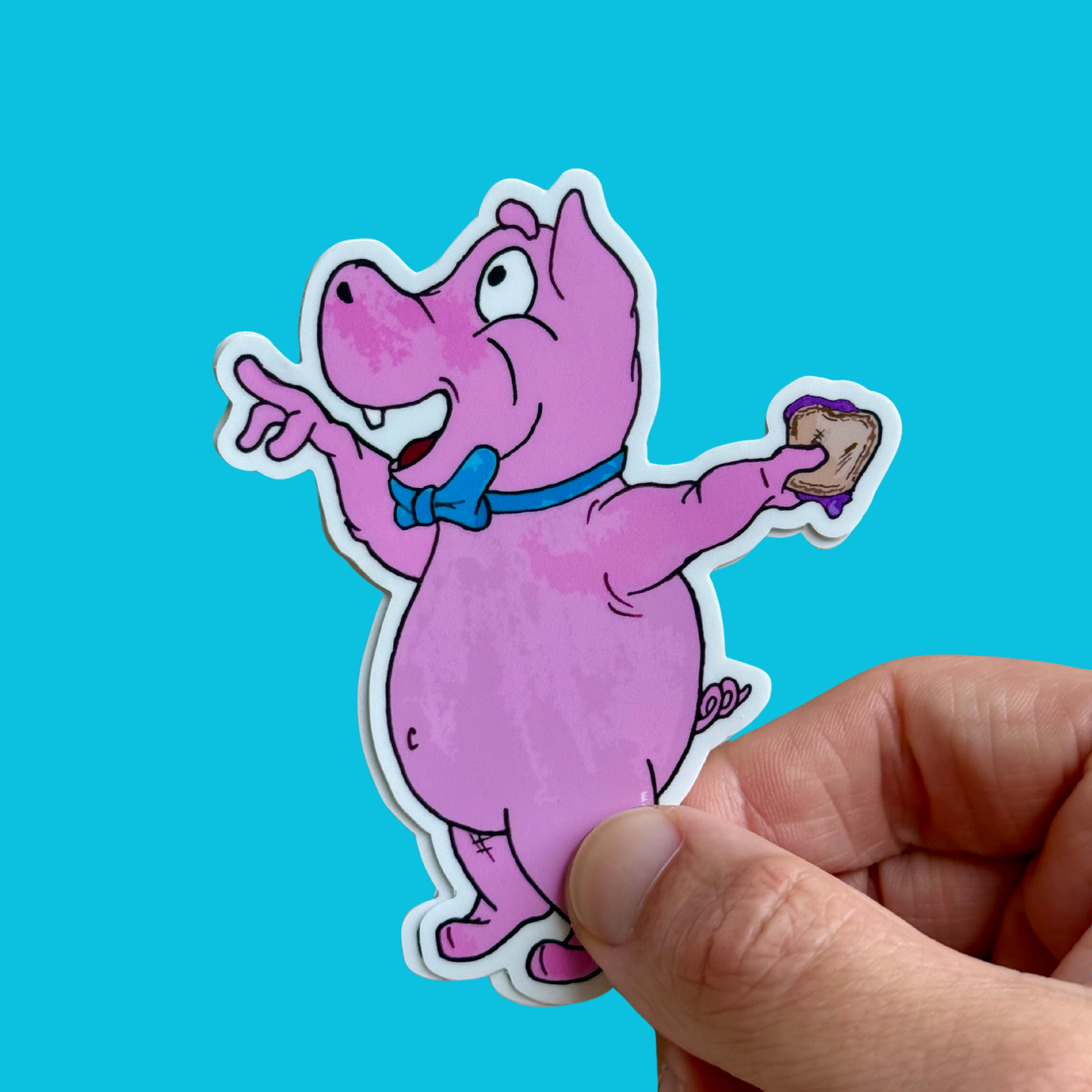 A die-cut sticker of a pink cartoon pig wearing a blue bow tie and holding a sandwich, held by a person's hand against a bright cyan background. A new character in the world of Prpl Up Pup.