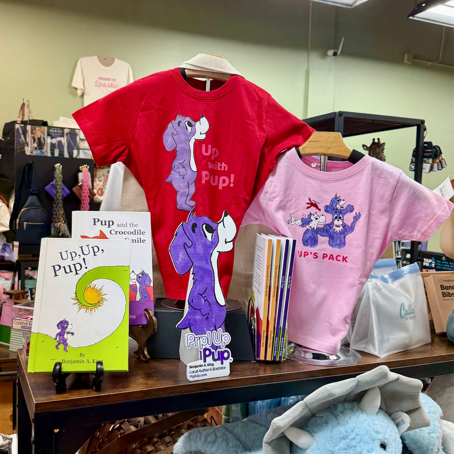 Display of children's t-shirts and Prpl Up Pup picture books with cartoon dog designs on a table. Find Prpl Up Pup children's brand at Woodland Cotton in Northport, Alabama.