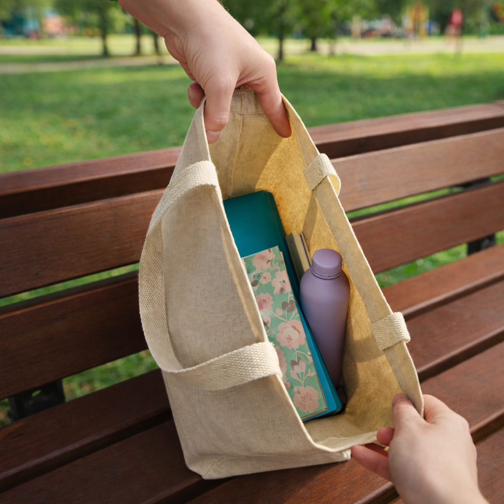 Person holding a Canvas Tote bag with a book and purple bottle on a wooden bench in a park. The Pup Love Tote is a mom's essential for every day moments.