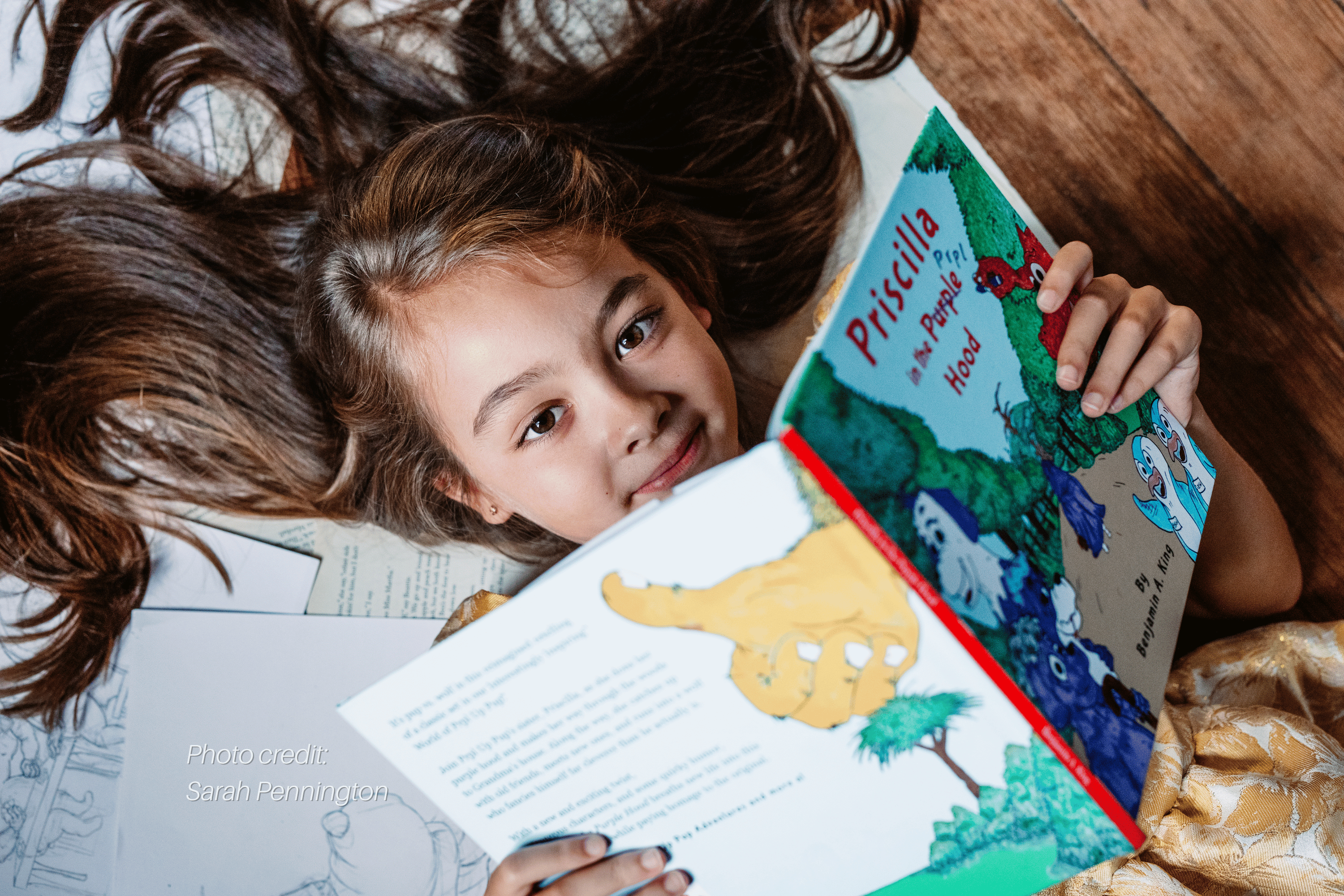 A young girl with long brown hair lies on a wooden floor reading a colorful children's book titled 'Priscilla and the People Who Flocked.'.